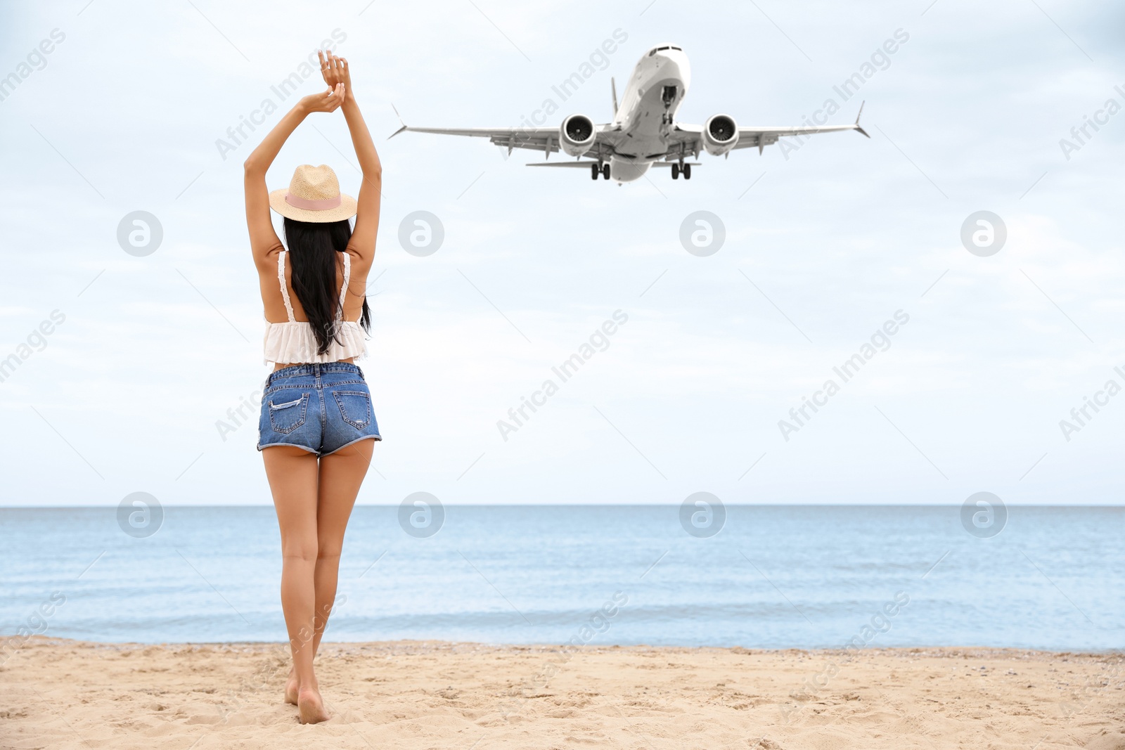 Image of Woman on sandy beach looking at airplane flying in sky, back view