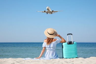 Image of Woman with suitcase on sandy beach looking at airplane flying in sky, back view