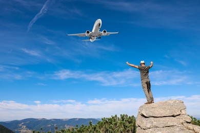 Image of Man on rocky peak looking at airplane flying in sky, back view