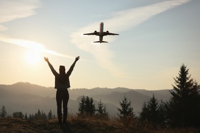 Image of Woman with raised arms in mountains looking at airplane flying in sky, back view