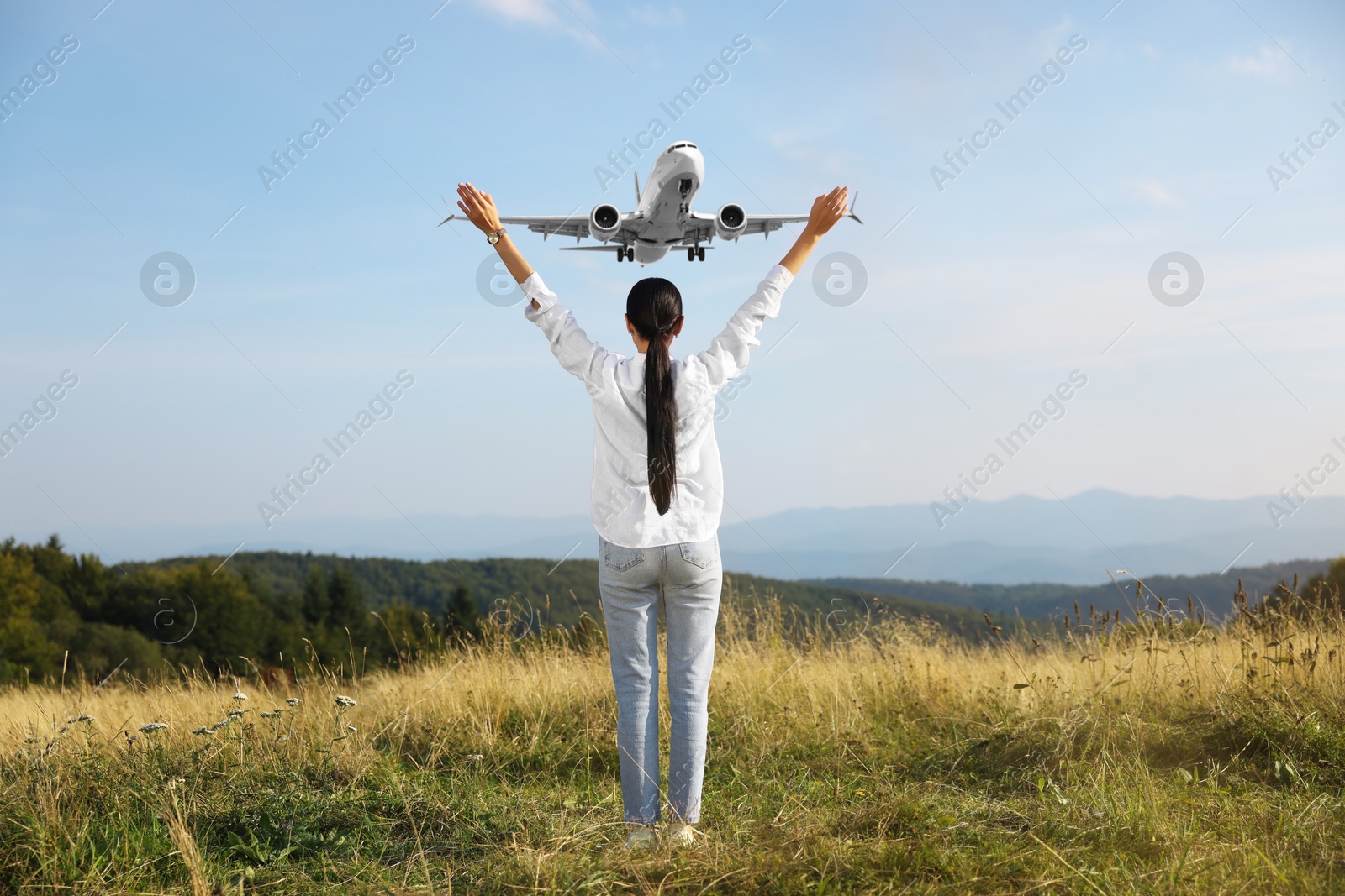 Woman with raised hands in mountains looking at airplane flying in sky, back view Image of Woman with raised hands in mountains looking at airplane flying in sky, back view