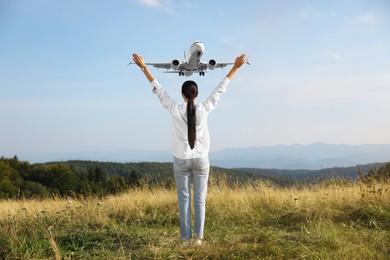 Image of Woman with raised hands in mountains looking at airplane flying in sky, back view