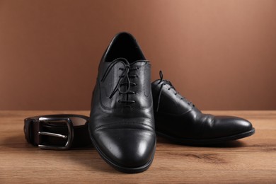 Photo of Pair of black leather shoes and belt on wooden surface against brown background, closeup