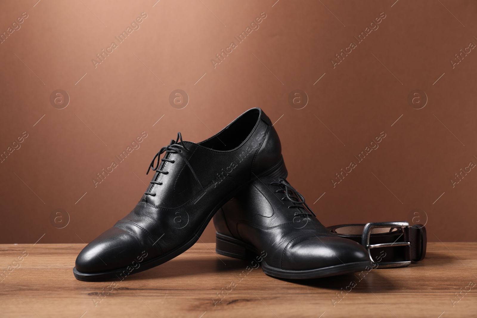 Pair of black leather shoes and belt on wooden surface against brown background Photo of Pair of black leather shoes and belt on wooden surface against brown background