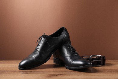 Photo of Pair of black leather shoes and belt on wooden surface against brown background
