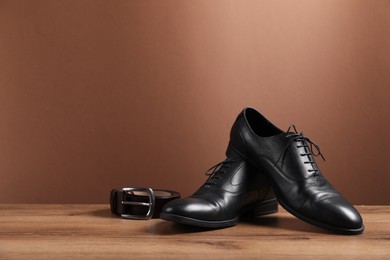 Photo of Pair of black leather shoes and belt on wooden surface against brown background, space for text