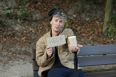 Photo of Homeless man with sign Help and paper cup begging for money on bench in park