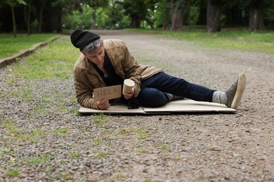 Photo of Homeless man with sign Help and paper cup lying on carton in park