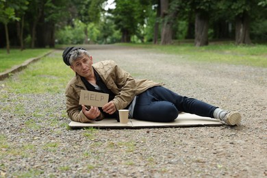 Photo of Homeless man with sign Help and paper cup lying on carton in park