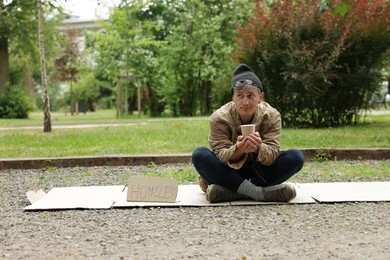 Photo of Poor man with sign Homeless and paper cup begging for money on carton in park