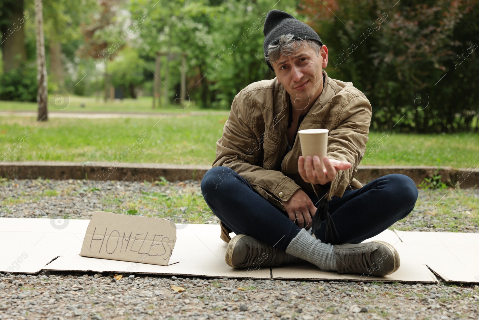 Poor man with sign Homeless and paper cup begging for money on carton in park Photo of Poor man with sign Homeless and paper cup begging for money on carton in park