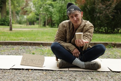 Photo of Poor man with sign Homeless and paper cup begging for money on carton in park