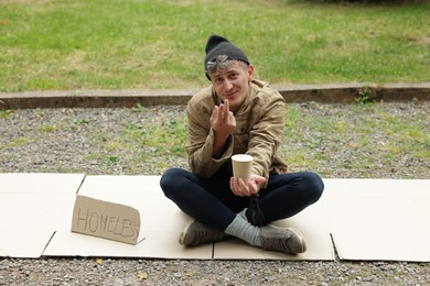 Photo of Poor man with sign Homeless and paper cup begging for money on carton in park