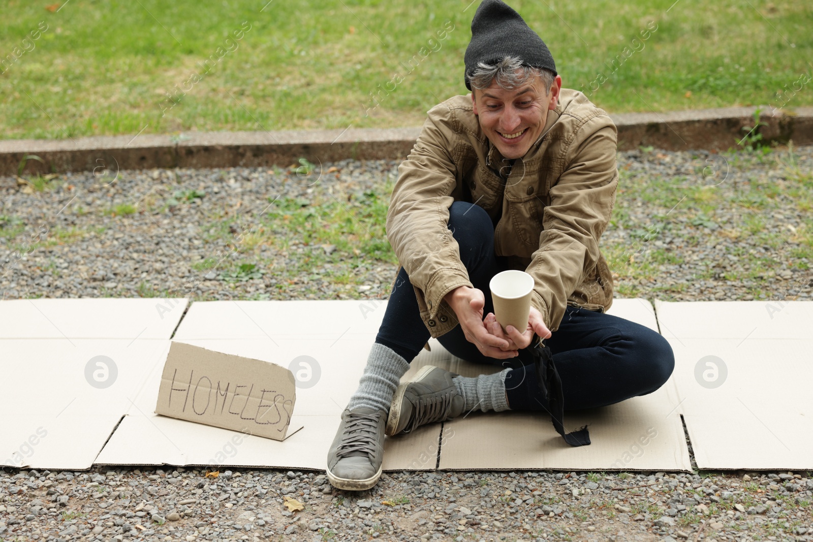 Poor man with sign Homeless and paper cup begging for money on carton in park Photo of Poor man with sign Homeless and paper cup begging for money on carton in park