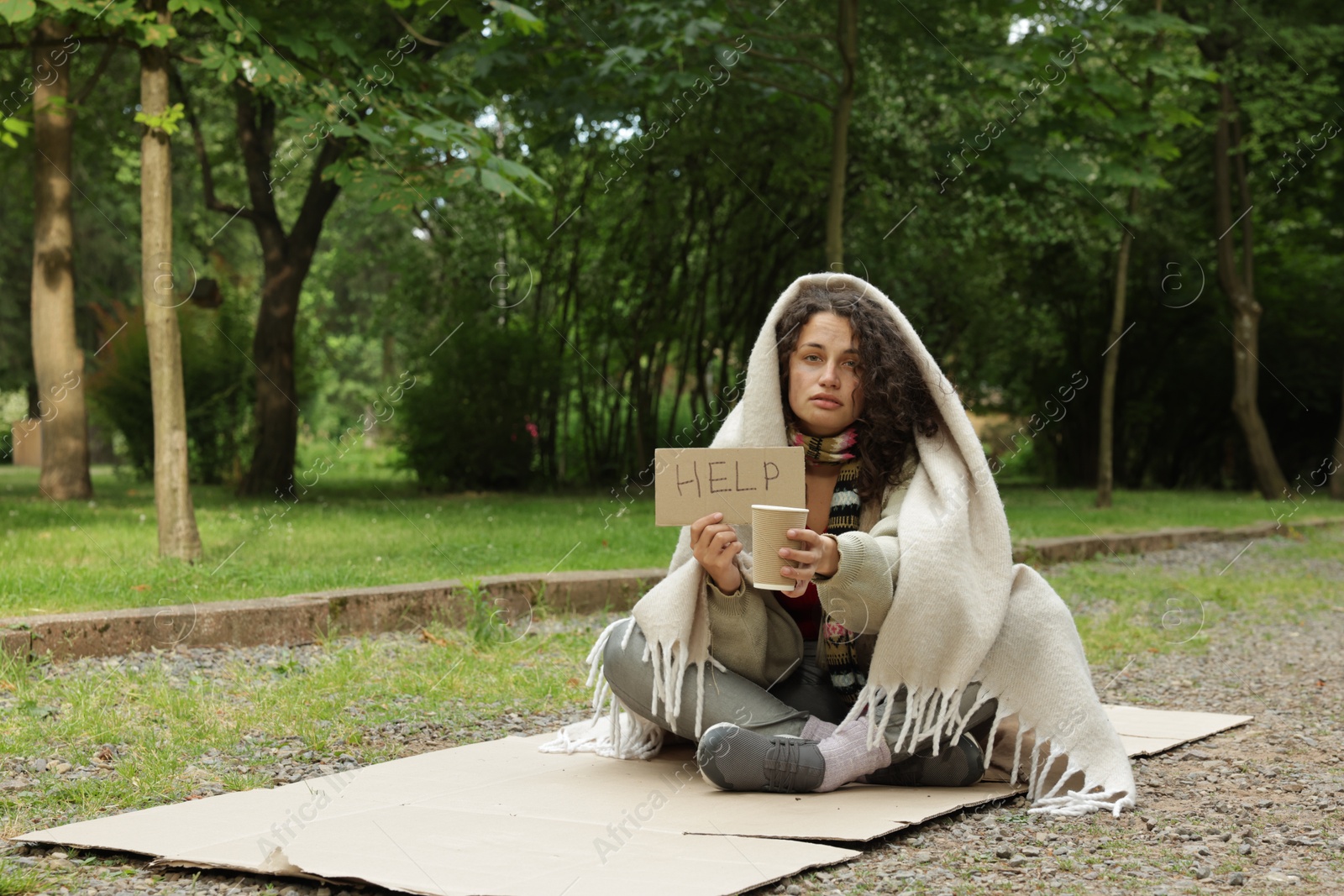 Homeless woman with sign Help and paper cup on carton in park, space for text Photo of Homeless woman with sign Help and paper cup on carton in park, space for text