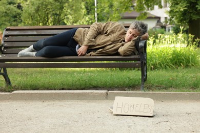 Photo of Poor man sleeping on bench and sign Homeless in park