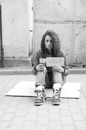 Photo of Homeless woman with sign Help and paper cups on carton outdoors. Black and white effect