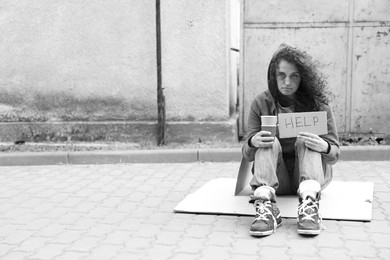 Photo of Homeless woman with sign Help and paper cups on carton outdoors, space for text. Black and white effect