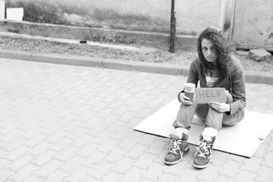 Photo of Homeless woman with sign Help and paper cups on carton outdoors, space for text. Black and white effect