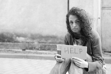 Photo of Homeless woman with sign Help and paper cups outdoors, space for text. Black and white effect
