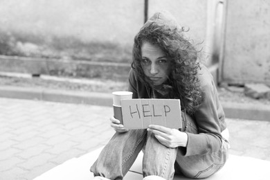 Photo of Homeless woman with sign Help and paper cups on carton outdoors, space for text. Black and white effect