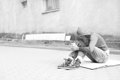 Photo of Homeless woman with paper cup on carton outdoors, space for text. Black and white effect