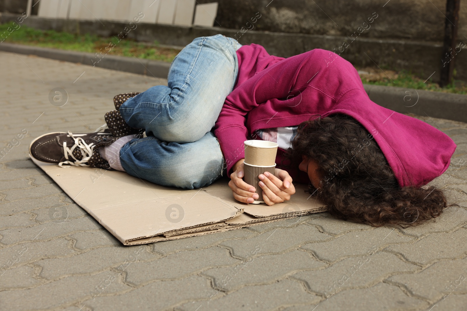 Homeless woman with paper cups lying on carton outdoors Photo of Homeless woman with paper cups lying on carton outdoors