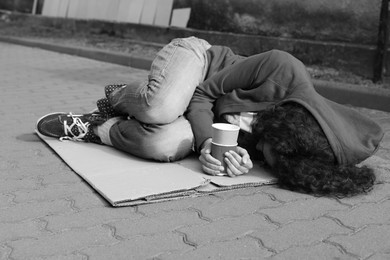 Photo of Homeless woman with paper cups lying on carton outdoors. Black and white effect
