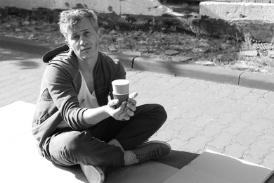 Photo of Homeless man with paper cups begging for money on carton outdoors, space for text. Black and white effect