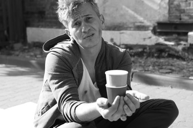 Photo of Homeless man with paper cups begging for money outdoors. Black and white effect