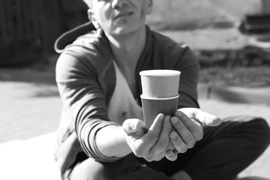 Photo of Homeless man with paper cups begging for money outdoors, closeup. Black and white effect