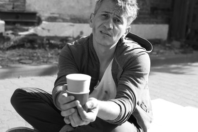 Photo of Homeless man with paper cups begging for money outdoors. Black and white effect