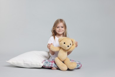 Photo of Cute little girl in pajamas with teddy bear and pillow on grey background
