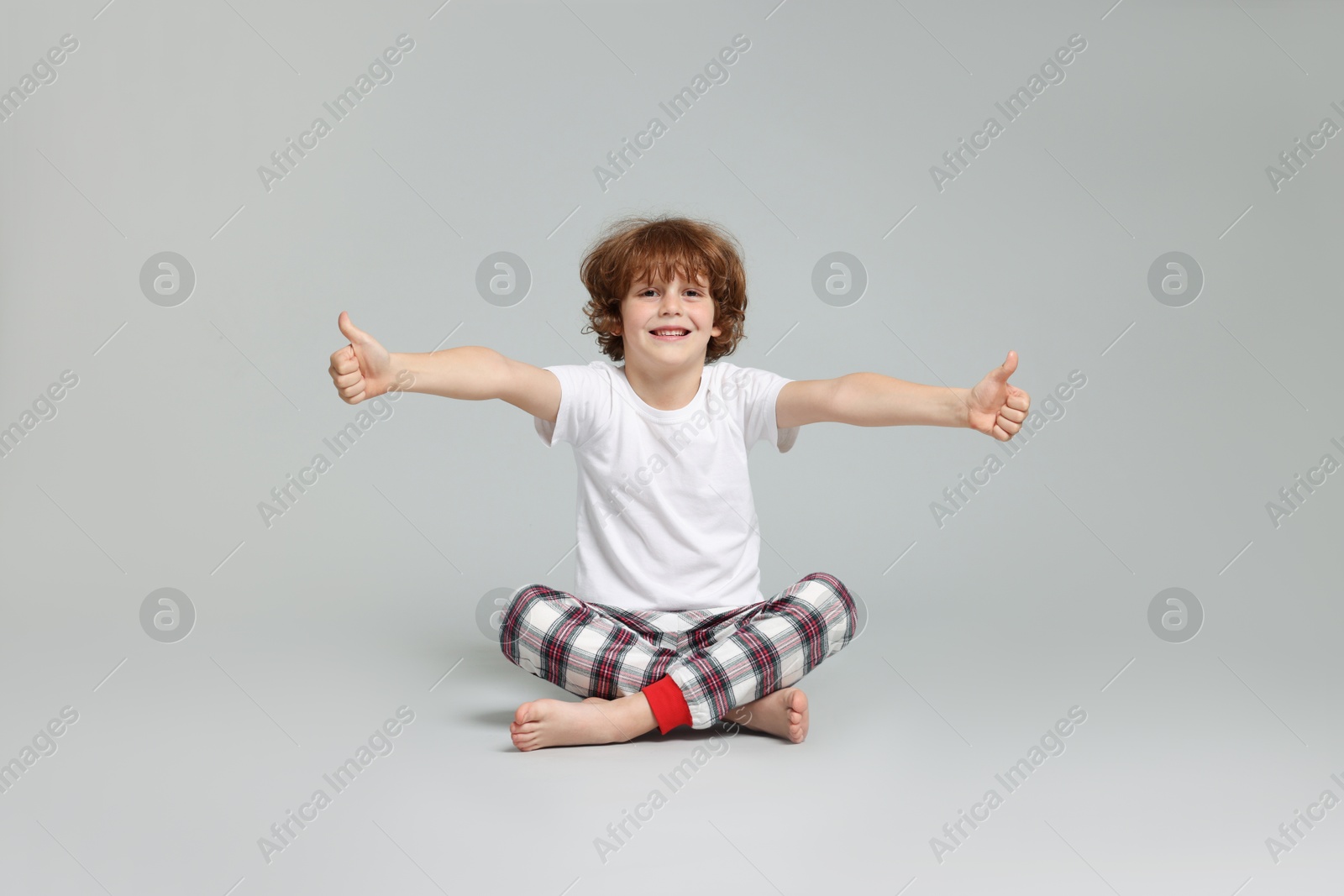 Photo of Cute little boy in pajamas showing thumbs ups on grey background
