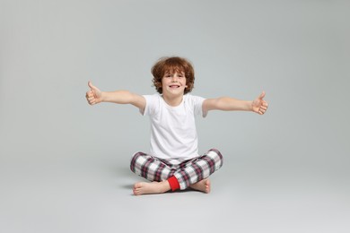 Photo of Cute little boy in pajamas showing thumbs ups on grey background