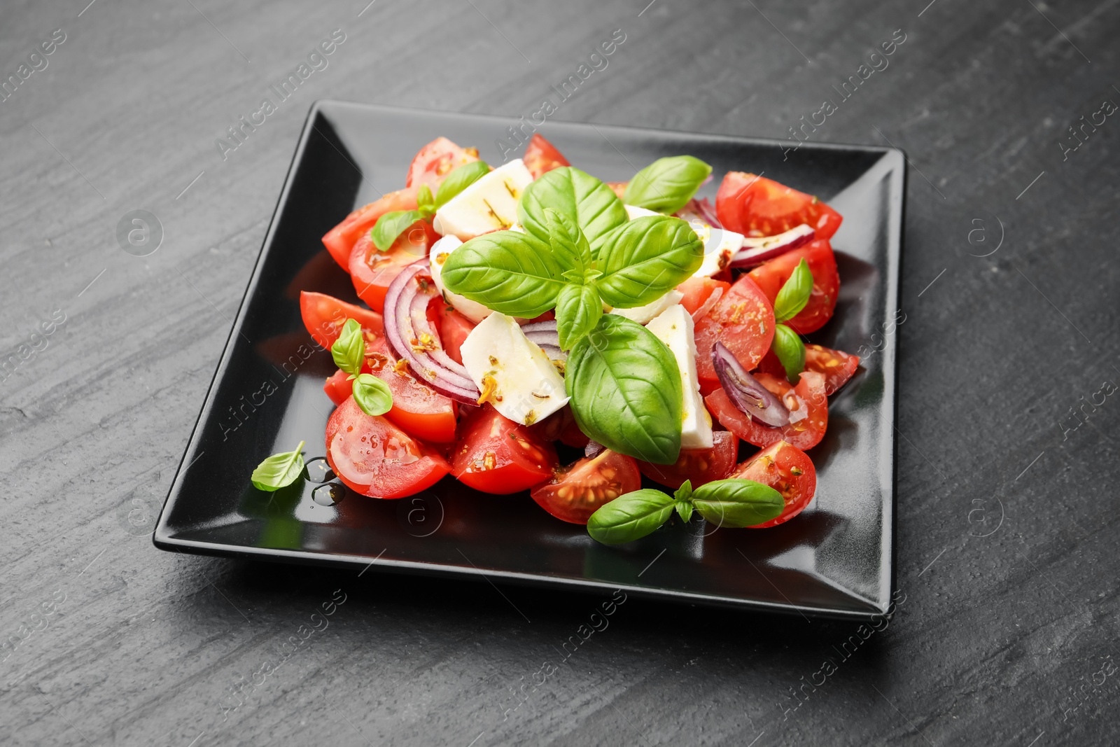 Tasty salad Caprese with mozzarella, tomatoes, basil, onion and spices on black table, closeup Photo of Tasty salad Caprese with mozzarella, tomatoes, basil, onion and spices on black table, closeup