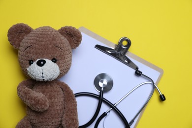 Photo of Pediatrics concept. Toy bear with stethoscope and clipboard on yellow background, top view