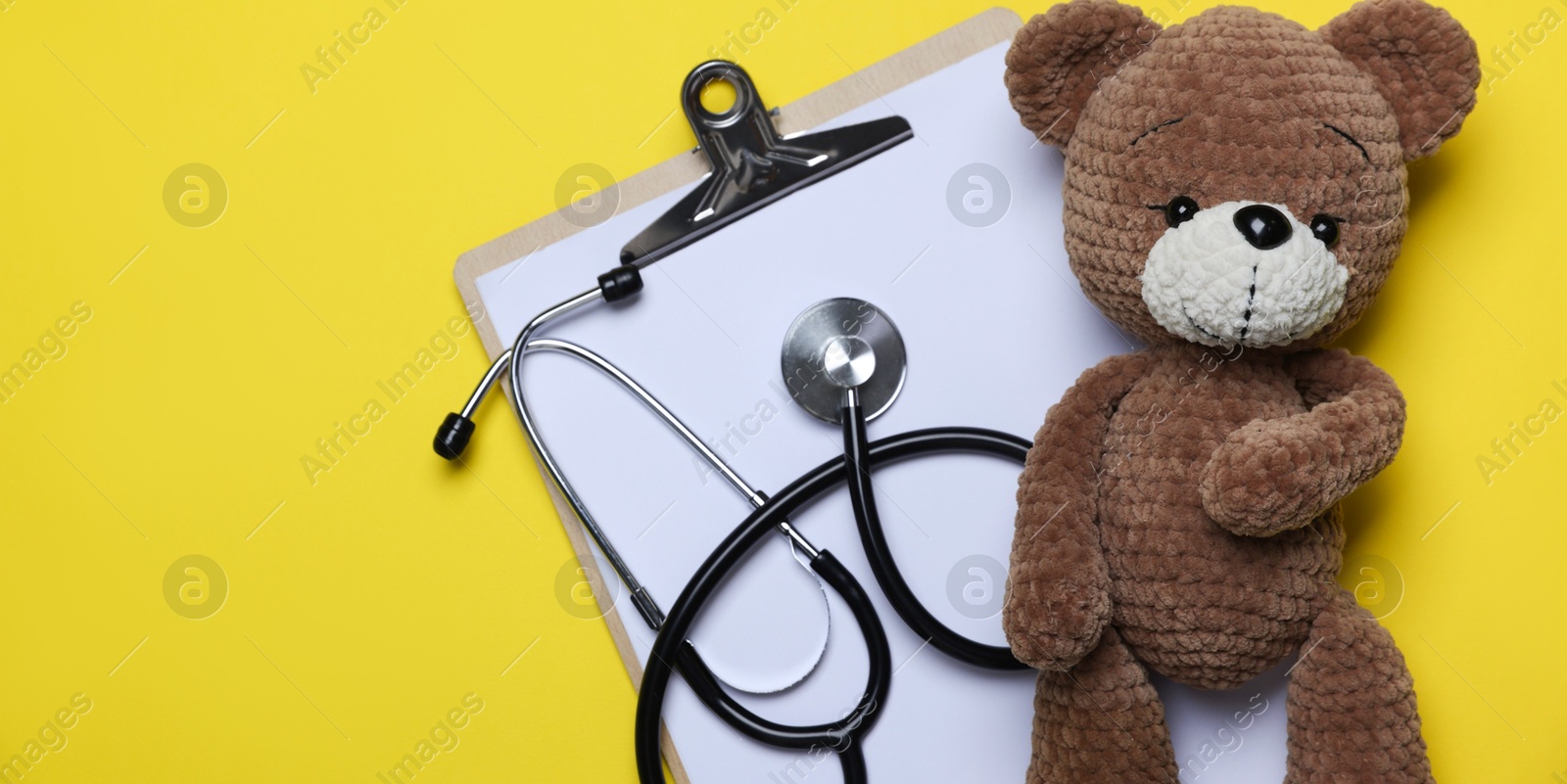 Photo of Pediatrics concept. Toy bear with stethoscope and clipboard on yellow background, top view. Space for text