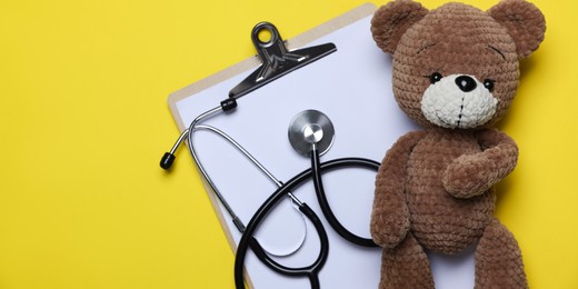 Photo of Pediatrics concept. Toy bear with stethoscope and clipboard on yellow background, top view. Space for text
