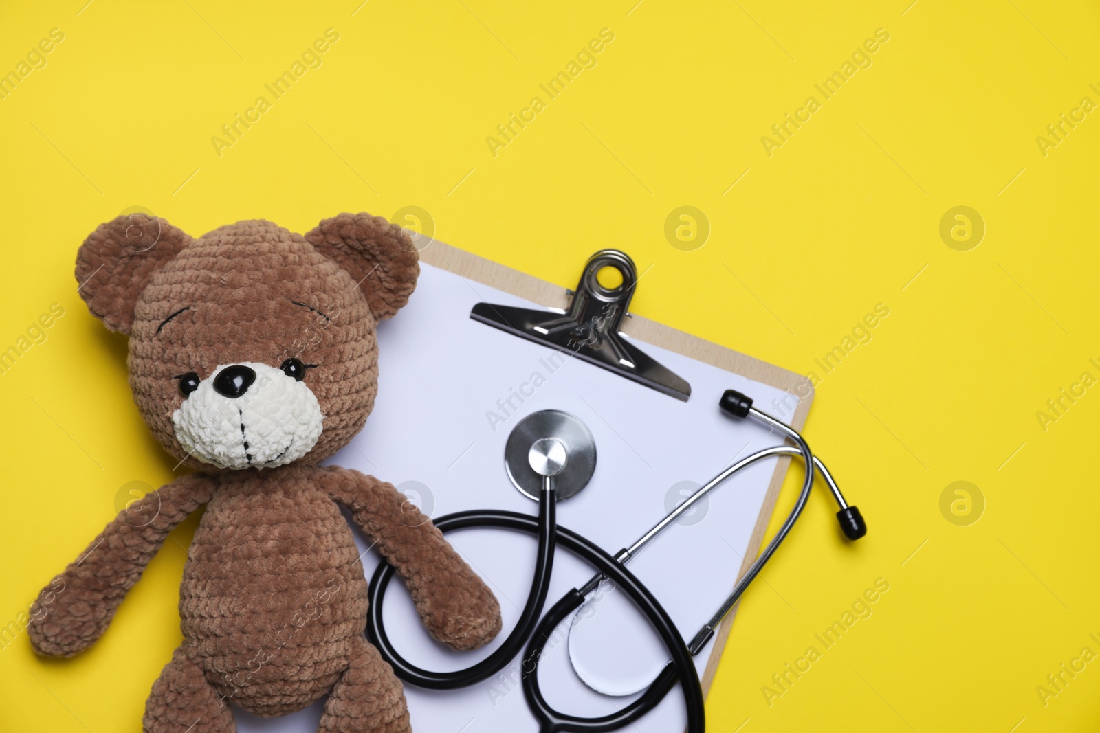 Photo of Pediatrics concept. Toy bear with stethoscope and clipboard on yellow background, top view