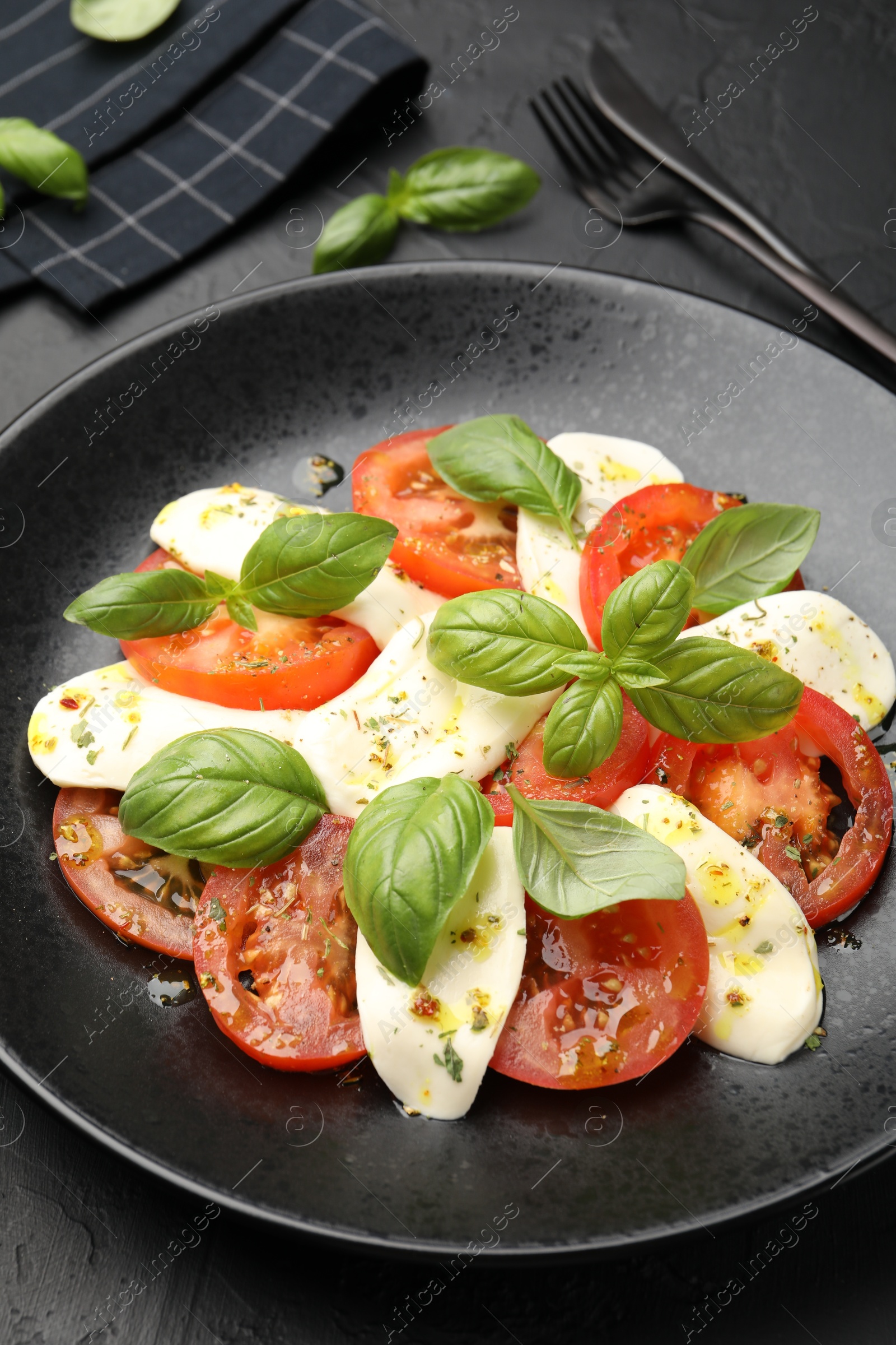 Photo of Tasty salad Caprese with mozzarella, tomatoes, basil and spices on black table, closeup