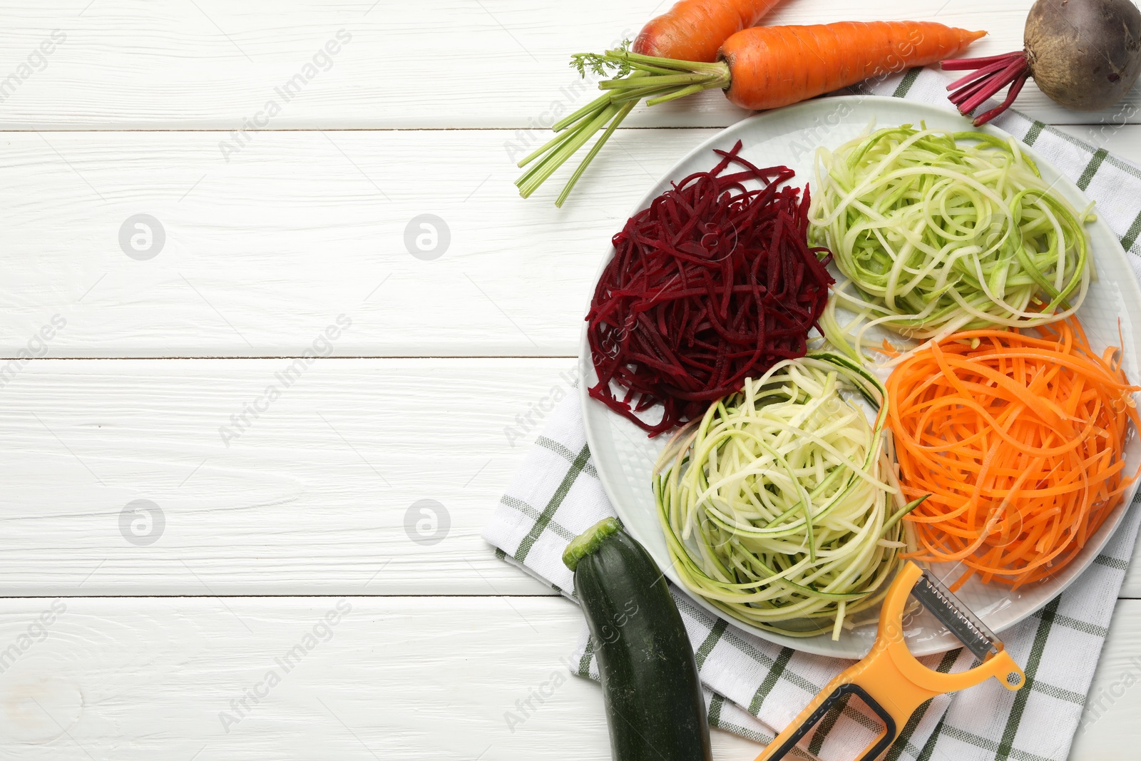 Photo of Vegetarian dish. Different vegetable noodles, fresh vegetables and peeler on white wooden table, flat lay. Space for text