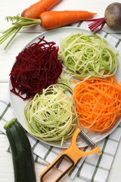 Photo of Different vegetable noodles, fresh vegetables and peeler on white wooden table, flat lay. Vegetarian dish