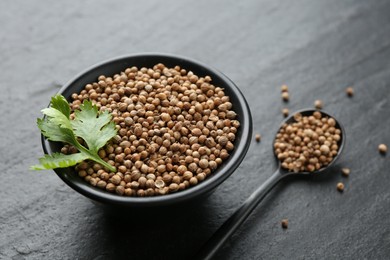 Photo of Coriander seeds in bowl, spoon and fresh cilantro leaves on dark textured table, closeup