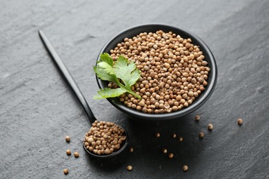 Photo of Coriander seeds in bowl, spoon and fresh cilantro leaves on dark textured table, closeup