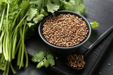 Photo of Coriander seeds in bowl, spoon and fresh cilantro leaves on dark textured table, closeup