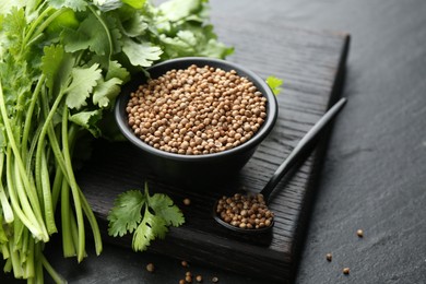 Photo of Coriander seeds in bowl, spoon and fresh cilantro leaves on dark textured table, closeup