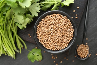 Photo of Coriander seeds in bowl, spoon and fresh cilantro leaves on dark textured table, flat lay