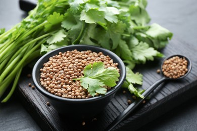 Photo of Coriander seeds in bowl, spoon and fresh cilantro leaves on dark table, closeup
