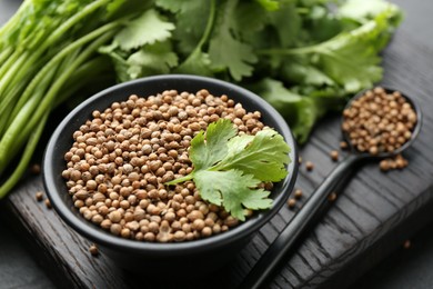 Photo of Coriander seeds in bowl, spoon and fresh cilantro leaves on dark table, closeup